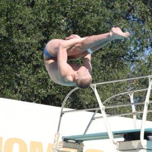 Ben Willett is upside-down in mid-flip during a dive. His legs are straight as he wraps his arms behind his knees. In the backdrop is a diving board and green tree foliage.
